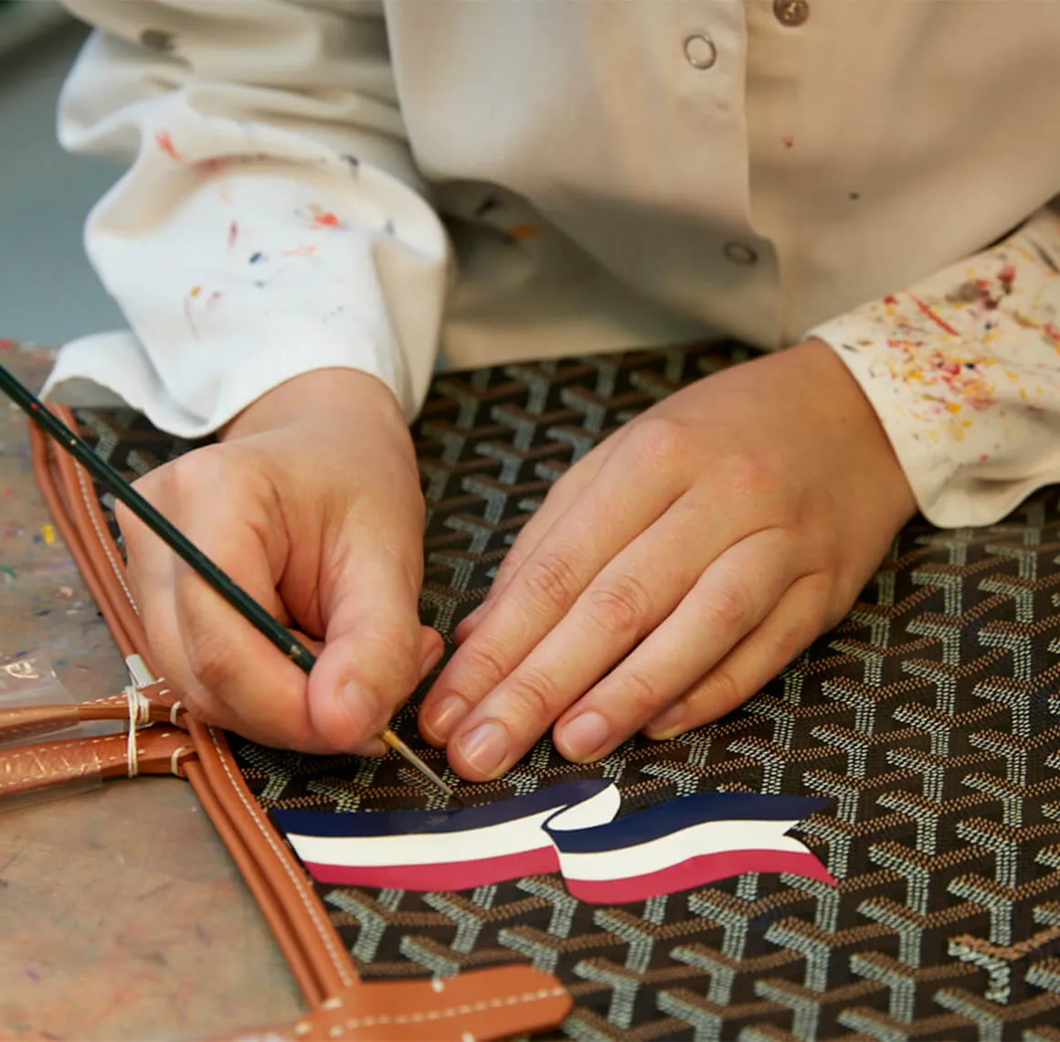 มือช่างใช้พู่กันเพนต์ลายสีแดง ขาว และน้ำเงินบนหีบ Goyard