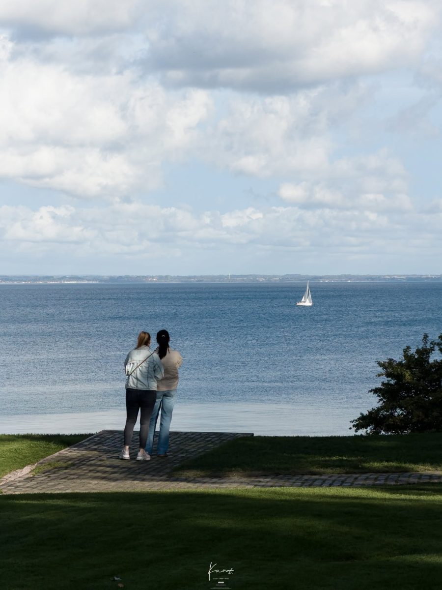 คนสองคนยืนมองทะเลช่องแคบ Øresund จากสนามหญ้าของพิพิธภัณฑ์ มีเรือใบอยู่ไกล ๆ