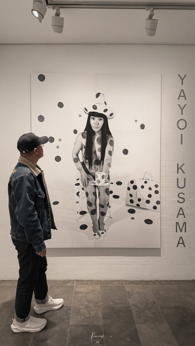 ผู้ชมยืนหน้าผนังลายจุดที่มีภาพและชื่อ Yayoi Kusama